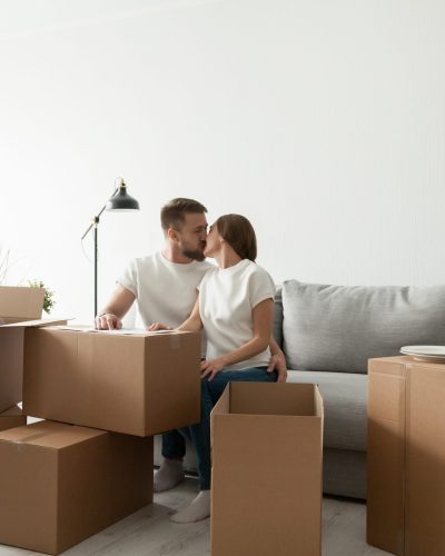 Young couple kissing sitting on sofa in living room with cardboard boxes on the floor, moving day in own home or relocation concept, happy homeowners real estate buyers starting new life together
